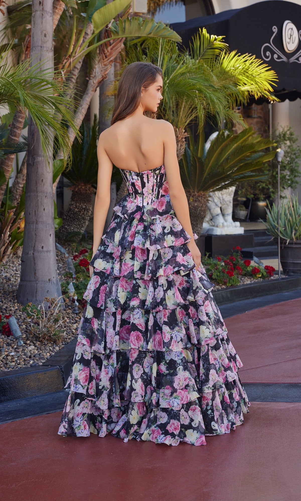 Outdoors, a woman faces away from the camera in the Nox Anabel Q1779 Long Prom Dress, a strapless corset gown with pink, purple, and black florals and chiffon tiered ruffle details.